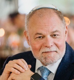 Rabbi David Levin, a white man with white hair and a white beard, wearing a black suit and blue tie, smiling and clasping his hands for the camera.