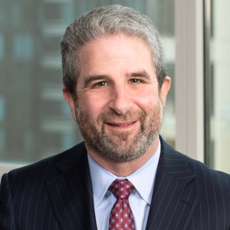 White man with gray hair and a black beard wearing a black suit and maroon tie