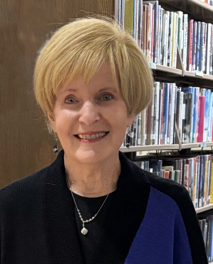 White woman with blonde hair wearing a black and blue sweater with books in the background